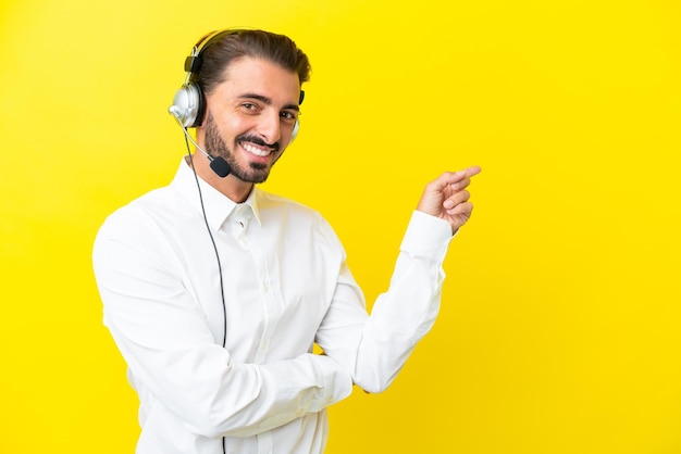 Telemarketer uomo caucasico che lavora con un auricolare isolato su sfondo giallo che punta il dito di lato