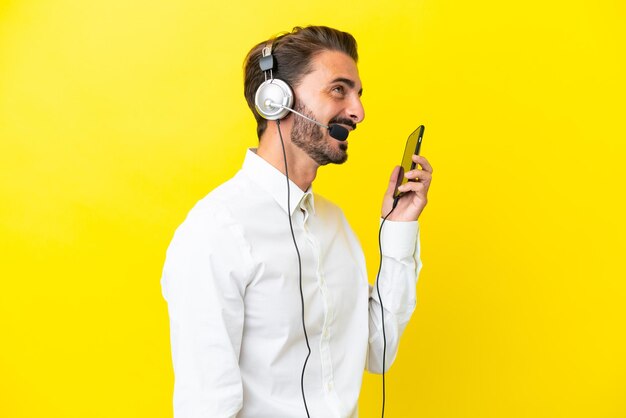 Telemarketer uomo caucasico che lavora con un auricolare isolato su sfondo giallo mantenendo una conversazione con il telefono cellulare