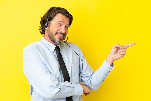 Telemarketer uomo olandese che lavora con un auricolare isolato su sfondo giallo che punta il dito di lato