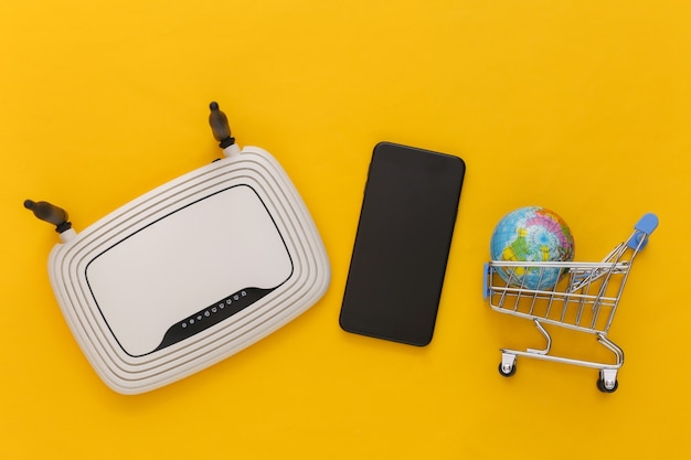 Tema dello shopping online. Carrello della spesa con globo, router wi-fi, smartphone su sfondo giallo. Vista dall'alto