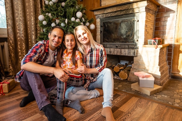 Tema Natale e Capodanno cerchio familiare. Giovane famiglia caucasica che si siede sul pavimento di legno a casa nel soggiorno vicino al camino Albero di Natale.