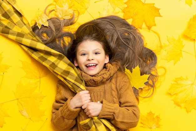 Tempo d'autunno Una bambina si trova su uno sfondo giallo tra le foglie d'acero cadute in autunno Un bambino in un caldo maglione lavorato a maglia e sciarpa