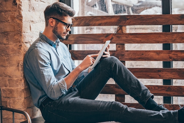 Tempo dei social media. Vista laterale di un bel giovane premuroso che lavora sul touchpad mentre è seduto al davanzale della finestra