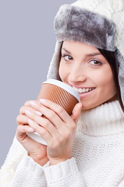 È tempo di bevande calde. Primo piano di giovani donne in abiti invernali caldi che bevono caffè e sorridono mentre sono in piedi su uno sfondo grigio