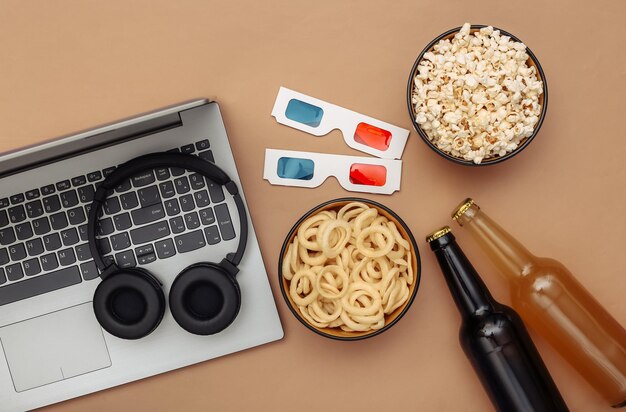 Tempo di intrattenimento. Ciotola per laptop e snack, cuffie, occhiali 3d, bottiglia di birra su sfondo marrone. Vista dall'alto