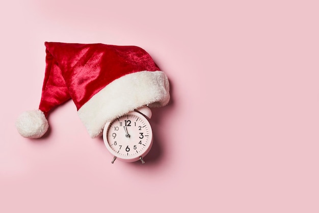 Tempo di NataleOrologio di Natale in un cappello o berretto di Babbo Natale su uno sfondo rosa La lancetta dei minuti a dodici o39 a Capodanno