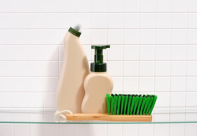 Tempo di pulizia e lavaggio Dispensatori di detergente beige e un pennello con spesse setole verdi su uno sfondo di piastrelle bianche