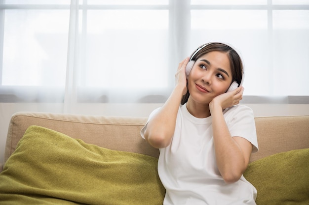 Tempo di relax sul divano. La bella giovane donna asiatica che si siede sul sofà sceglie la musica dallo smartphone riposa e ascolta la canzone con le cuffie. Stile di vita in soggiorno a casa al mattino.