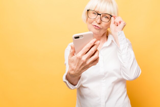 Tempo di selfie! Donna invecchiata positiva che sorride mentre usando il telefono isolato sopra priorità bassa gialla.