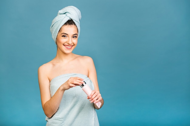 Tempo di Spa! Ritratto di bella donna sorridente in asciugamano sul corpo e sulla sua testa che tiene la lozione per il corpo isolata sopra priorità bassa blu.