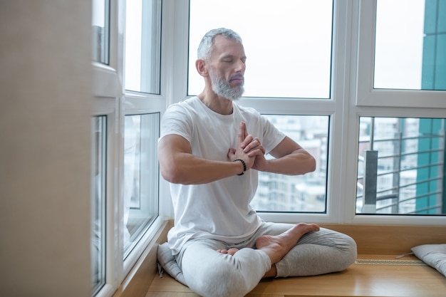 Tempo di yoga. Uomo dai capelli grigi con una maglietta bianca che fa yoga e sembra concentrato