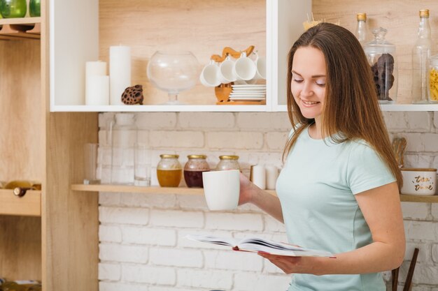 Tempo libero. Cucina di casa. Intimità serale. Bella giovane donna con la tazza della bevanda e del libro preferiti.