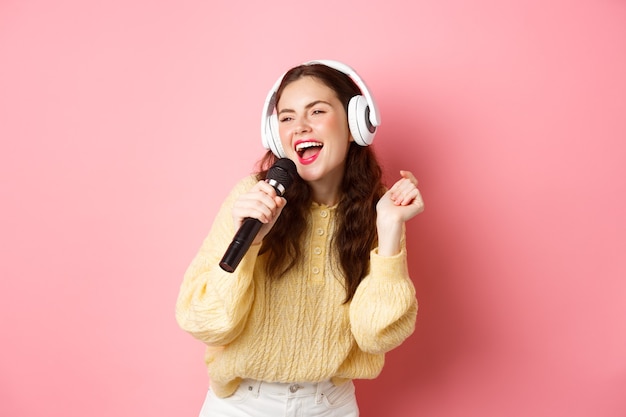 Tempo libero e stile di vita felice ragazza bruna divertendosi cantando karaoke in cuffie tenendo il microfono e fissando da parte lo schermo in piedi sopra la parete rosa