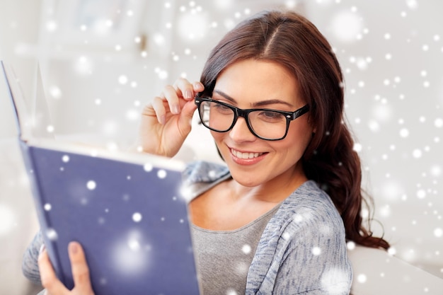 tempo libero, educazione, visione, inverno e concetto di persone - giovane donna con gli occhiali che legge il libro a casa sulla neve