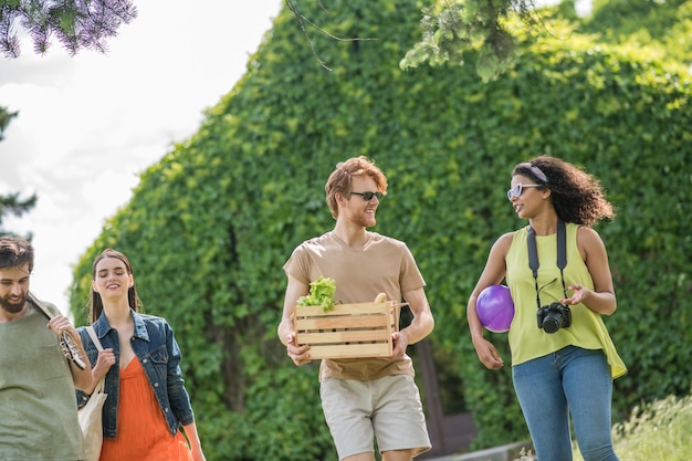 Tempo libero estivo. Giovani ottimisti energici due ragazze e ragazzi che comunicano andando a fare un picnic nella natura in una bella giornata estiva