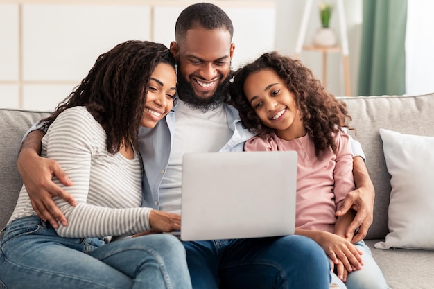 Tempo libero insieme. Ritratto di genitori neri positivi e figlia piccola che usano il laptop insieme a casa, guardando le foto di famiglia, scegliendo regali nel negozio online seduto sul divano in soggiorno