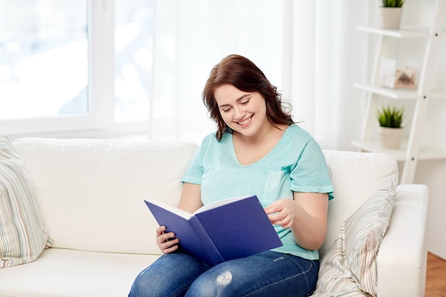 tempo libero, letteratura e concetto di persone - sorridente giovane donna di taglia più grande libro di lettura a casa