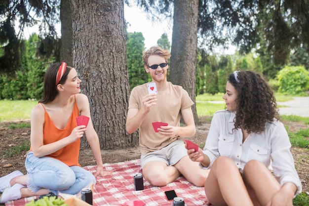 Tempo libero nella natura. Tre giovani amici allegri in abiti casual che giocano a carte seduti su una coperta sotto l'albero al picnic