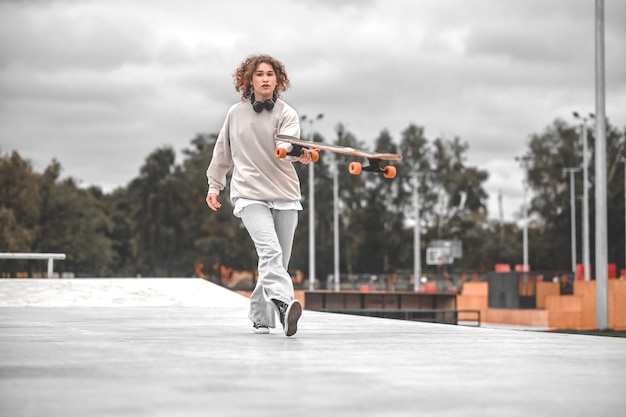 Tempo libero, skatepark. Ragazza con le cuffie sul collo in felpa e jeans che cammina sullo skatepark con lo skeetboard che trascorre il tempo libero all'aperto