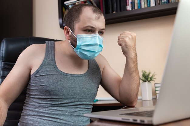 Tempo libero utilizzando laptop. Un giovane ragazzo barbuto con maschera facciale in abiti da casa utilizza il laptop