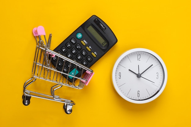 Tempo per lo shopping. Carrello per supermercati con calcolatrice, orologio su una superficie gialla. Minimalismo. Vista dall'alto