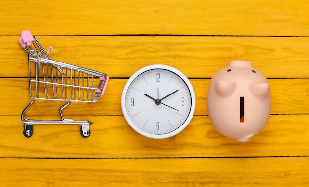 Tempo per lo shopping. Carrello per supermercato con salvadanaio, orologio su una superficie di legno gialla. Minimalismo. Vista dall'alto