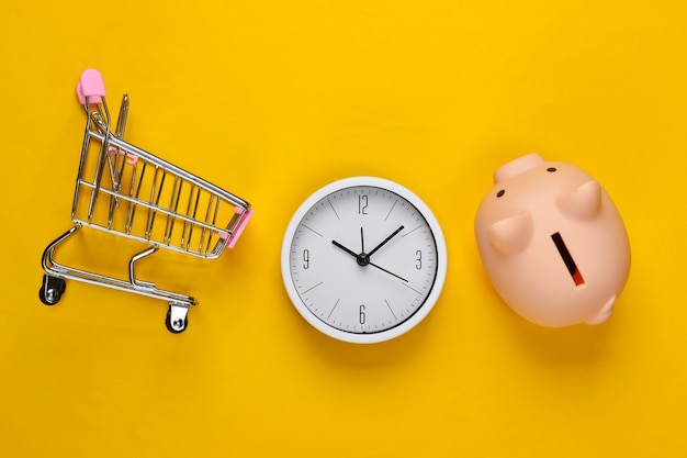 Tempo per lo shopping. Carrello per supermercato con salvadanaio, orologio su uno sfondo giallo. Minimalismo. Vista dall'alto