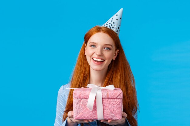 Tempo per presente Adolescente rossa allegra, studentessa universitaria festeggia il suo compleanno, con in mano un grazioso regalo incartato rosa e indossa un cappello da b-day, sorridendo con gioia in piedi su sfondo blu