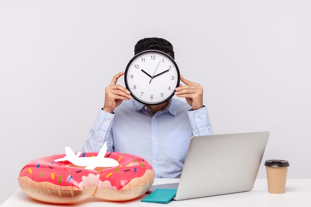 Tempo per riposare Uomo sconosciuto che nasconde la faccia dietro l'orologio seduto nel posto di lavoro dell'ufficio con passaporto ad anello di gomma e aeroplano di carta sul dipendente della scrivania che chiede una pausa relax e vacanze di viaggio in studio