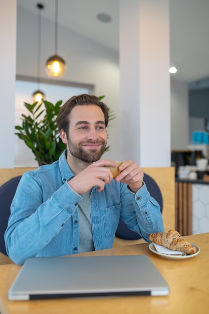 Tempo per te stesso. Felice giovane uomo barbuto che tiene la tazza seduto al tavolo con il laptop in un caffè che riposa sorridendo di lato