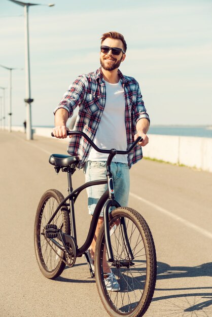 Tempo per un ciclo. Bel giovane uomo sorridente che cammina lungo una strada con la sua bicicletta