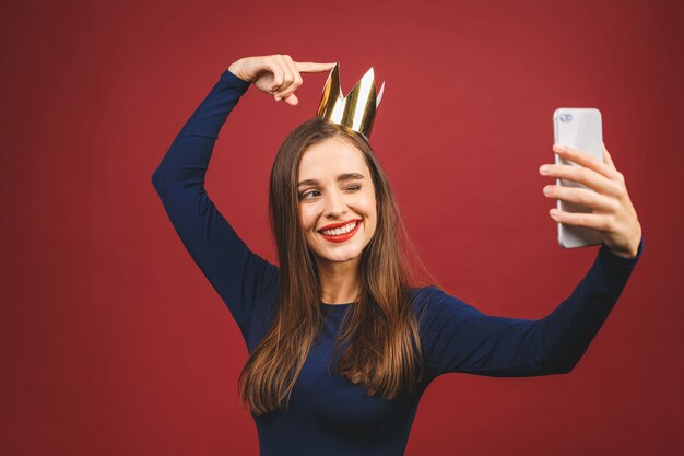 Tempo selfie! Ritratto con copyspace posto vuoto di fiduciosa orgogliosa giovane donna arrogante con corona d'oro sulla sua testa isolata su sfondo rosso.