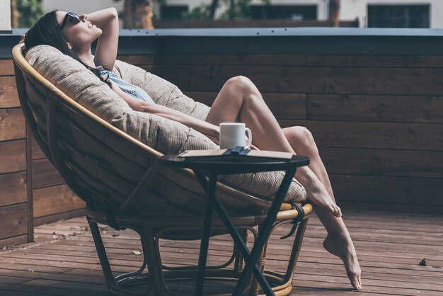 Tempo spensierato all'aperto. Bella giovane donna che si rilassa su una sedia grande e comoda sulla terrazza della sua casa all'aperto