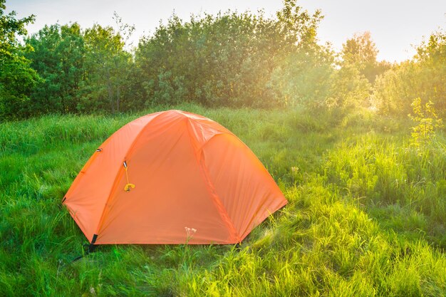 Tenda arancione che si accampa al tramonto nella foresta e nel campo di erba verde e nei raggi del sole