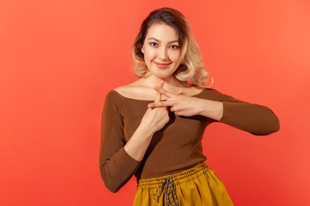 Tendenze Internet. Ritratto di affascinante donna felice in camicetta marrone incrociando le dita per fare il segno dell'hashtag e guardando la fotocamera con un sorriso. Studio indoor girato isolato su sfondo rosso