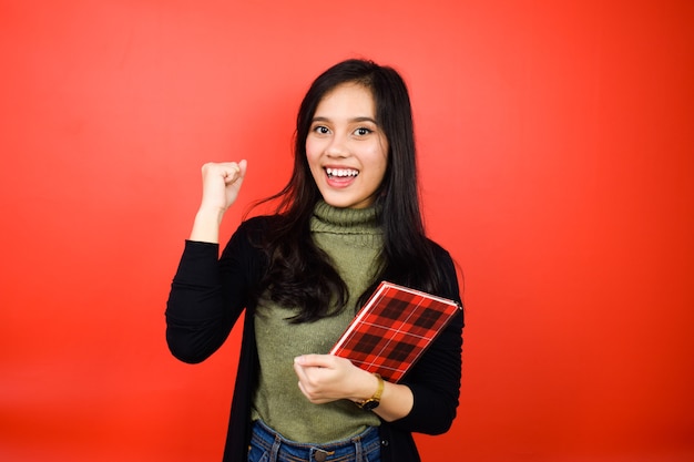 Tenendo il libro e sorridendo di belle donne asiatiche che usano un maglione nero con sfondo rosso isolato