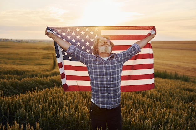 Tenendo in mano la bandiera degli Stati Uniti Uomo patriottico senior elegante con capelli grigi e barba sul campo agricolo