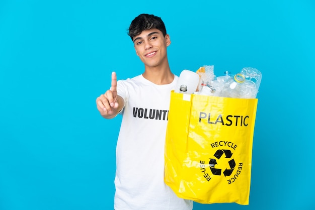 Tenendo una borsa piena di bottiglie di plastica da riciclare su blu mostrando e alzando un dito