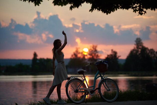 Tenera donna in abito in posa con una bicicletta d'epoca vicino al fiume sullo sfondo di un tramonto ammaliante