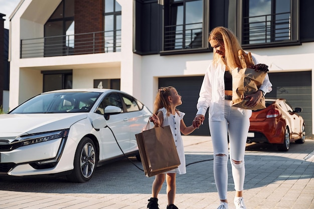 Tenere le borse mentre il veicolo è in carica Giovane donna con la sua piccola figlia è con la loro auto elettrica all'aperto