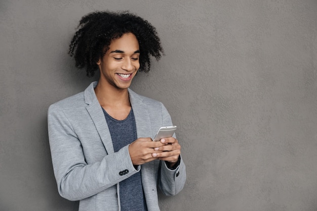 Tenersi in contatto. Allegro giovane africano che tiene in mano uno smartphone e lo guarda con un sorriso mentre si trova su uno sfondo grigio