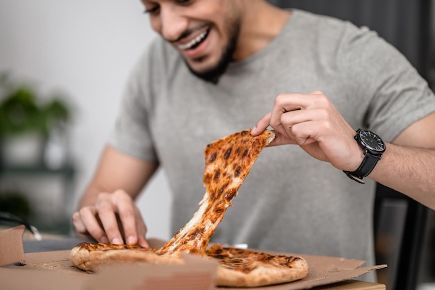 Tentazione. Uomo barbuto sorridente con un sorriso a trentadue denti che strappa la pizza in porzioni tenendo le dita seduto al tavolo