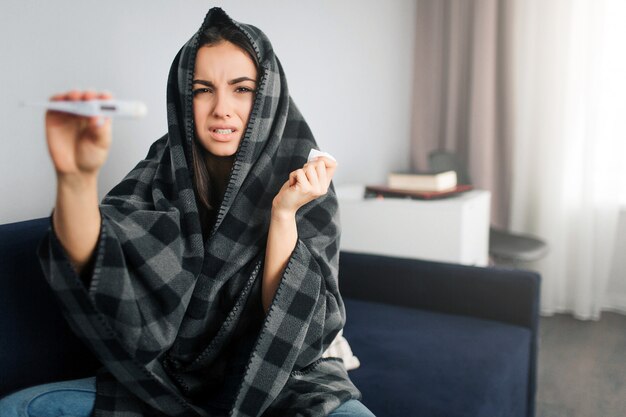 Termometro malato della stretta della giovane donna a disposizione. Lo mostra alla telecamera. Il modello ha la temperatura. Si copre di coperta.