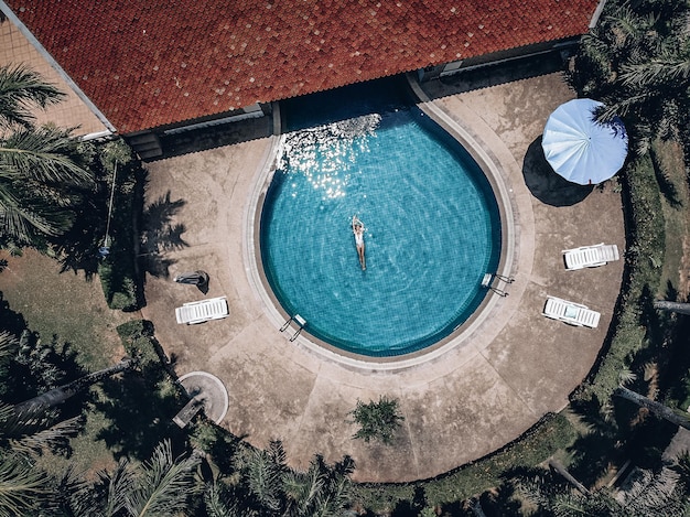 Territorio dell'hotel tropicale, bella donna galleggia sul retro nella piscina rotonda blu, macchie nell'acqua blu. Tegola in terracotta, palme, vista aerea