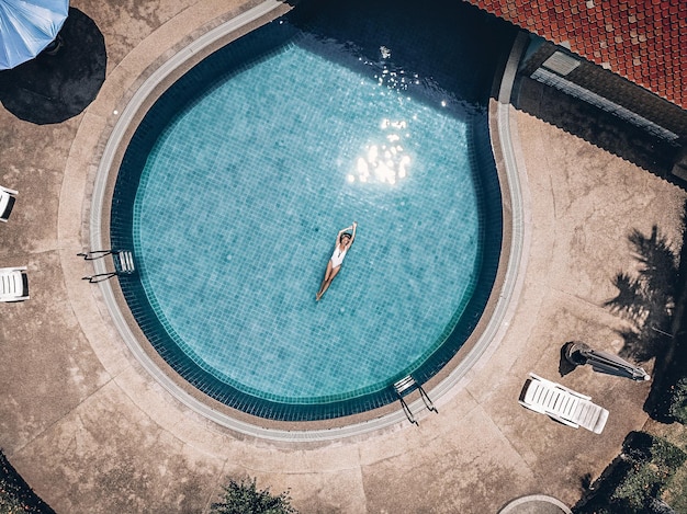 Territorio dell'hotel tropicale, bella donna galleggia sul retro nella piscina rotonda blu, macchie nell'acqua blu. Tegola in terracotta, palme, vista aerea