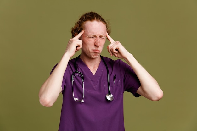 Teso con gli occhi chiusi che mettono le dita sulla fronte giovane medico maschio che indossa l'uniforme con uno stetoscopio isolato su sfondo verde