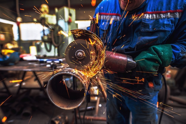 Tessuto maschio lavoratore taglio tubo metallico con una smerigliatrice elettrica in officina.