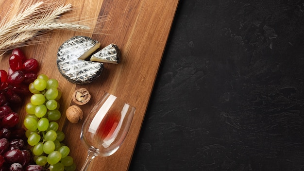 Testa di formaggio, grappolo d'uva, noci e bicchiere di vino su tavola di legno e sfondo nero. Vista dall'alto con copia spazio.