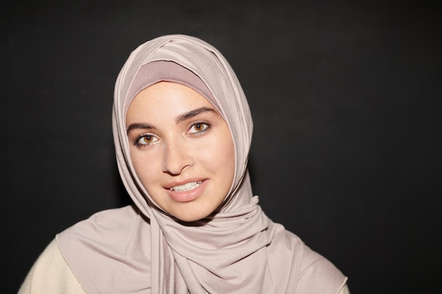 Testa e spalle ritratto di bella ragazza musulmana che indossa l'hijab in piedi contro il muro nero backg