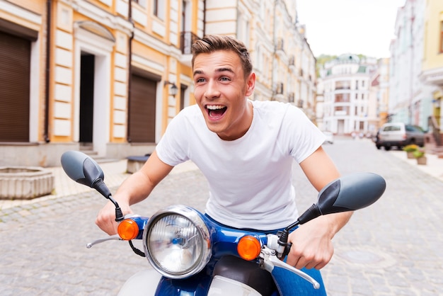 Testare il suo nuovo scooter. Felice giovane in sella a uno scooter lungo la strada e tenendo la bocca aperta